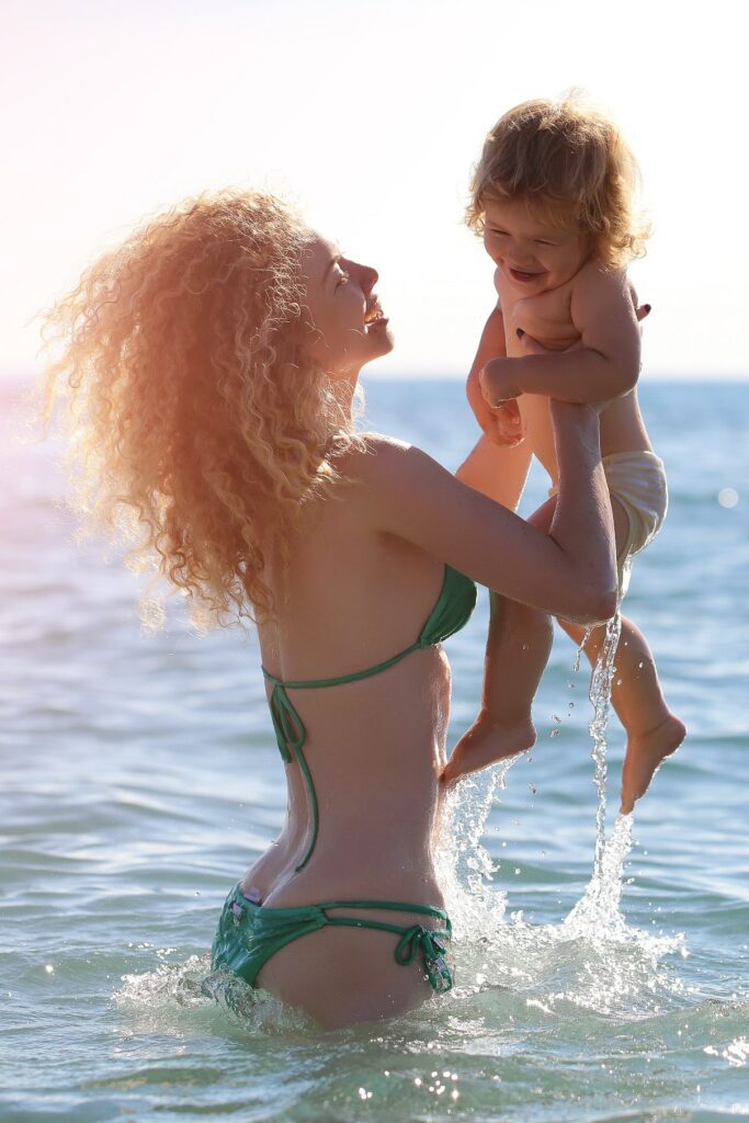 Woman in bikini with a flat belly after mommy makeover standing in the ocean holding up a toddler.
